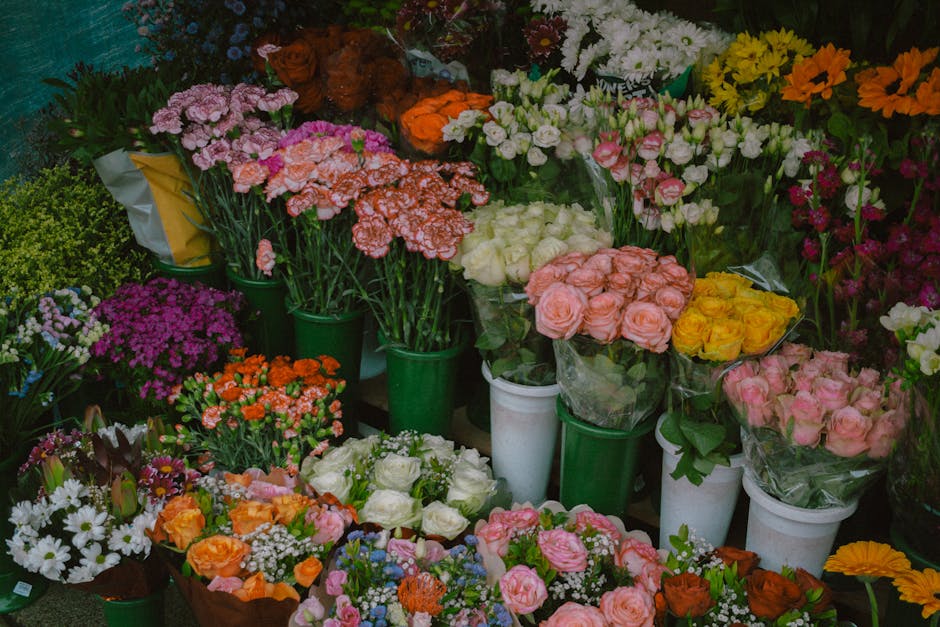 A vibrant display of fresh flowers arranged in various bouquets and potted plants. The collection includes pink roses, yellow roses, white roses, orange carnations, purple and white daisies, and clust