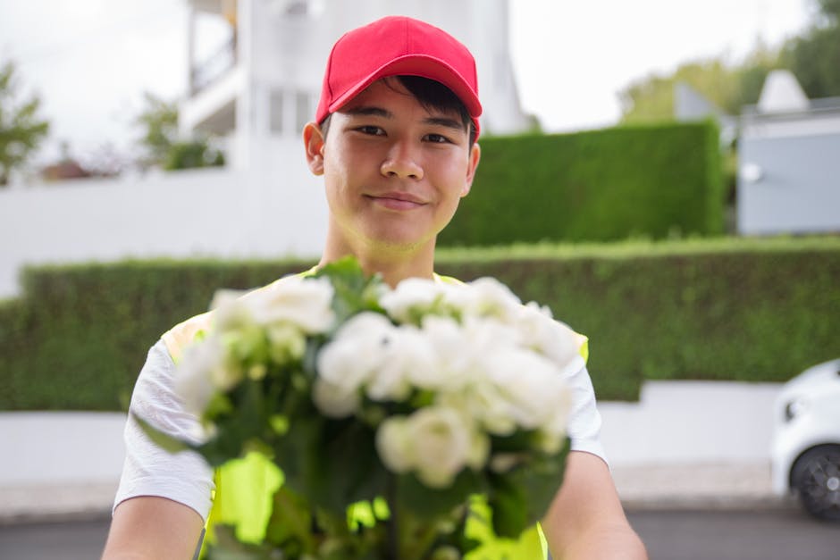 A delivery person wearing a red cap and neon yellow vest holds out a bouquet of fresh white flowers, including roses and peonies, wrapped in clear and green cellophane. The arrangement is lush with vi