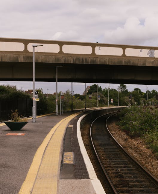 The image depicts a quiet, outdoor train platform with a curved railway track running alongside it. The platform features a yellow safety line and a tactile paving strip near the edge, with a small bl