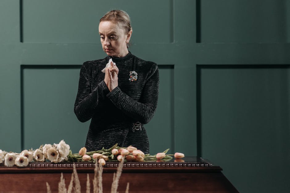 A woman dressed in a black textured dress with a floral brooch stands solemnly behind a dark wooden table set against a muted green wall. In front of her, there is a floral arrangement featuring white