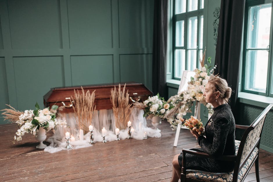 A woman seated in a floral arrangement exhibit or memorial setting, holding a bouquet of peach and pink flowers. She is dressed in a dark, textured outfit and has her hair styled in an elegant updo. I