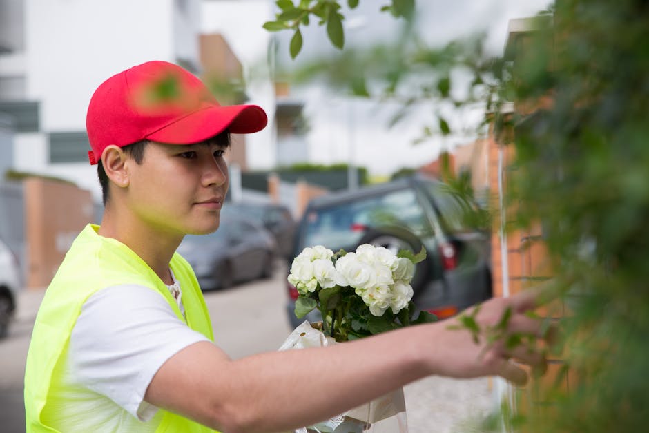 A young male florist wearing a red cap, white t-shirt, and neon yellow vest arranging a bouquet of fresh white hydrangeas wrapped in white paper. He is outdoors on a residential street, standing next