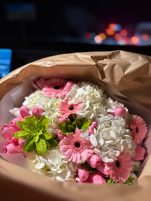A floral bouquet featuring white hydrangeas and pink gerbera daisies with dark centers, accented by bright green foliage. The flowers are arranged in a compact, rounded bouquet wrapped in light brown